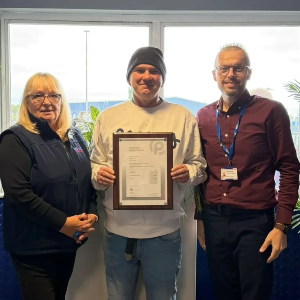 Michael holds his Record of Achievement certificate for completing his Level 2 Apprenticeship with Distinction, flanked by two NIC colleagues in an office setting.